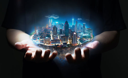 Digital composite image of hand holding illuminated cityscape against black background