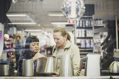 Saleswoman showing kettle to female customer in store seen through glass window