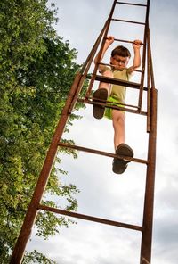 Low angle portrait of happy boy climbing ladder against sky