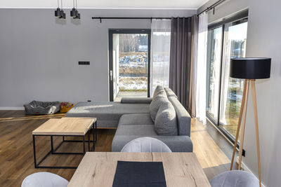 A modern and simple living room in the house with a gray sofa, large patio windows and tables.