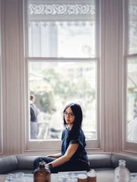 Asian woman in eyeglasses sitting beside big bay window in restaurant overlooking courtyard.