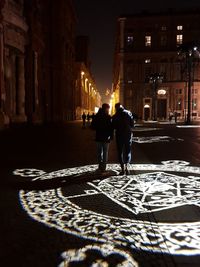 Rear view of man and woman walking on illuminated street at night