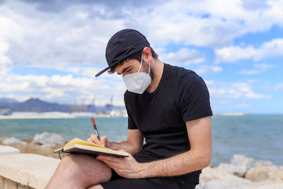 Male author in face mask and cap sitting with crossed legs on fence while taking note in diary behind ocean