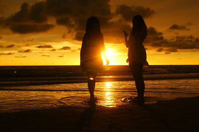 Silhouette female friends at beach against orange sky