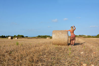 Rear view of woman standing by hay bales at field against sky
