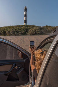 Blonde woman taking a photo her mobile phone the car to a lighthouse on a blue sky day