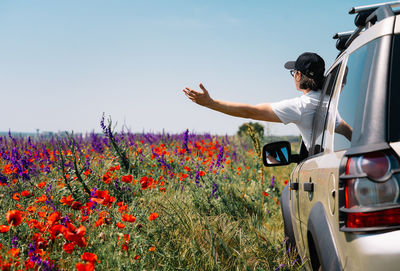 Driver an his car by scenic view of flowering wild plants on field against sky
