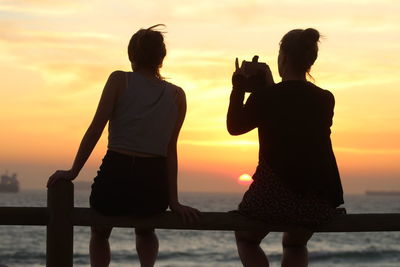 Rear view of woman photographing through smart phone while sitting with friend on railing during sunset