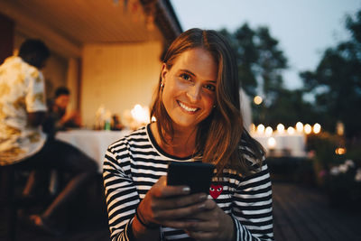 Portrait of smiling woman holding smart phone while friends in background during dinner party