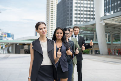 Portrait of smiling young colleagues showing thumbs up while standing on footpath against buildings in city