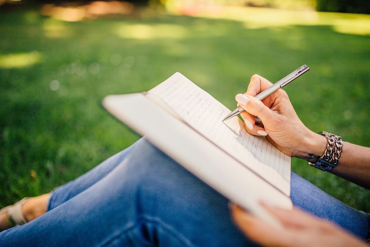 grass, adult, one person, relaxation, sitting, casual clothing, activity, leisure activity, publication, women, lifestyles, writing, book, nature, handwriting, plant, day, summer, hand, pen, reading, holding, selective focus, outdoors, paper, young adult, green, wireless technology, communication, limb, park, lawn