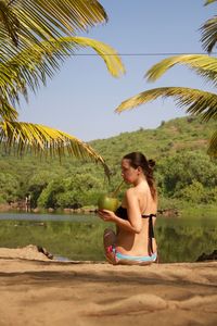 Rear view of woman drinking coconut while sitting against lake