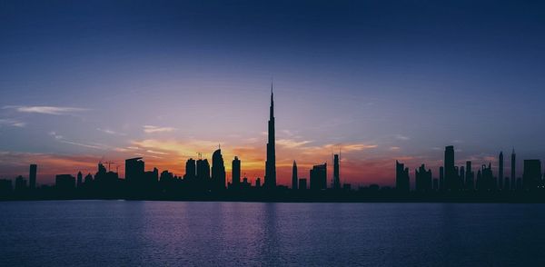 Silhouette buildings in front of sea against sky during sunset