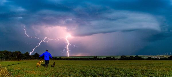 Man walking with dogs on grassy field against sky during thunderstorm