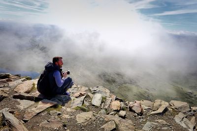 Side view of male hiker with camera sitting on mountain during foggy weather