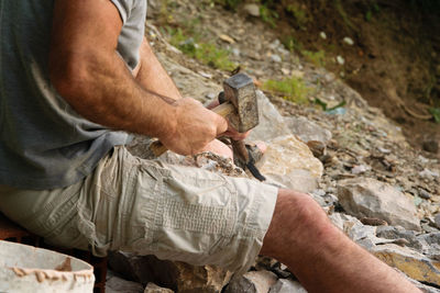 Midsection of man breaking rocks while sitting in forest