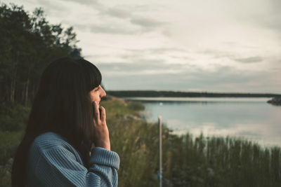 Woman talking on mobile phone while standing by lake against sky