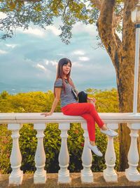 Young woman sitting on railing against trees