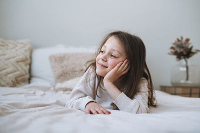 Cuty funny thinking little girl with long hair in home clothes lies on bed at home