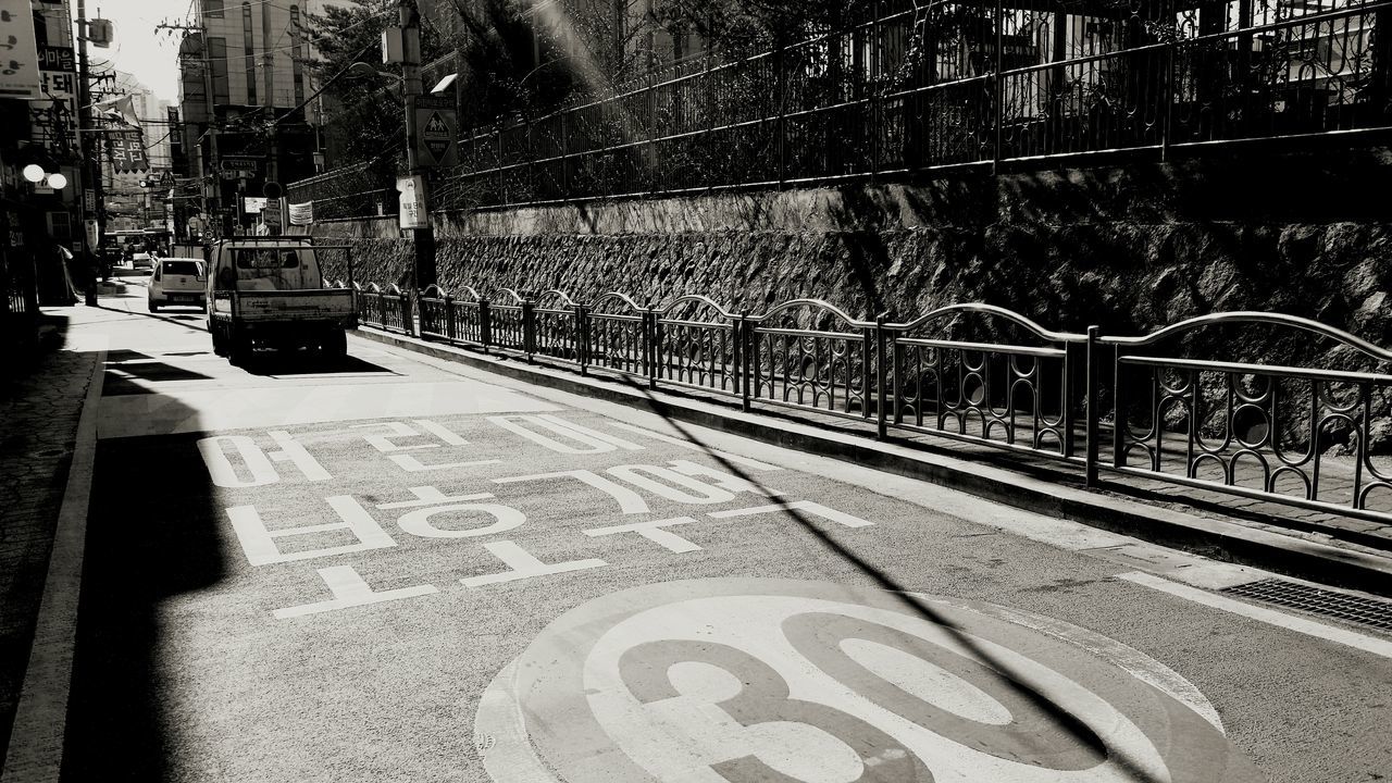 transportation, text, street, western script, road, communication, city, graffiti, fence, road marking, sunlight, shadow, land vehicle, railing, built structure, sidewalk, outdoors, empty, day, bicycle