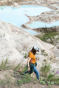 Young woman traveler in cap running on sand of clay quarry with blue water, landscape view hike