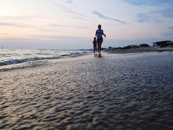 Mother and son running on beach against sky during sunset