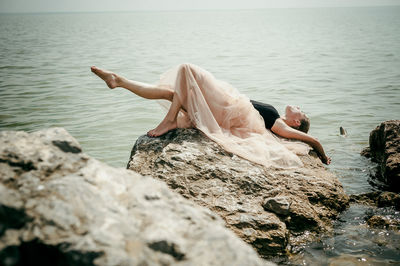 Young woman sitting on rock by sea against sky