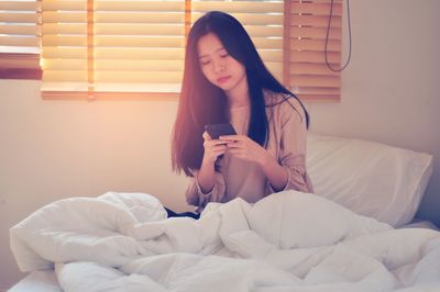 Young woman using mobile phone while sitting on bed at home