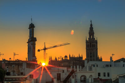 Cranes amidst buildings in city against sky during sunset