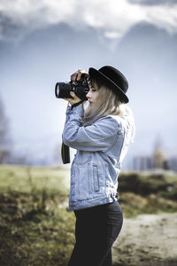 Side view of young woman photographing through camera against sky