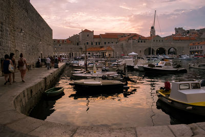 Sailboats moored on canal amidst buildings in city against sky during sunset