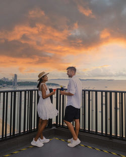 Rear view of woman with arms outstretched standing by railing against sky during sunset