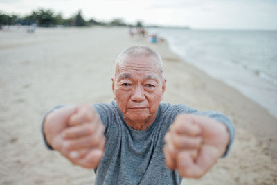 Portrait of senior man exercising while standing at beach against sky during sunset