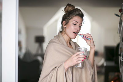 Young woman brushing teeth while holding glass of water