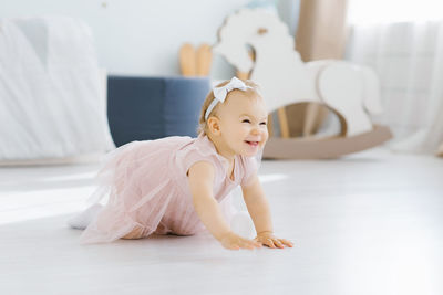 A pretty blue eyed blonde one year old in a pink dress crawls on all fours in the nursery 