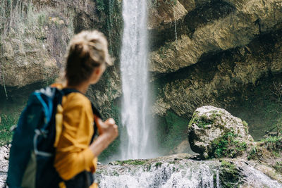 An unrecognizable defocused woman from behind with a backpack stands near a waterfall in a canyon.