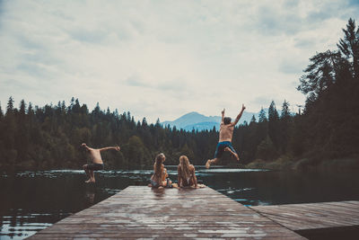 People jumping on pier over lake against sky