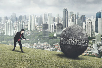 Businessman pulling rock with persistence text on hill against cityscape