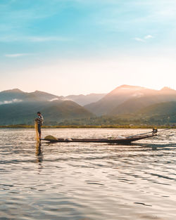 Man fishing in lake while standing on boat against mountains and sky