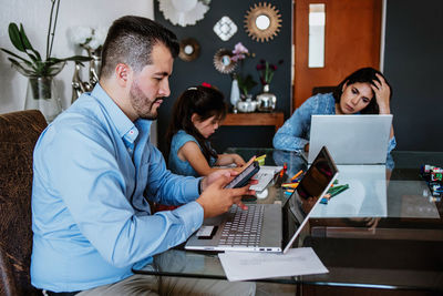 Busy exhausted young ethnic parents working remotely on laptops sitting at table at home near cute little daughter doing homework assignment