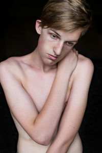 Shirtless young fashion model hugging self against black background