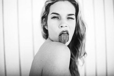 Portrait of young shirtless woman sticking out tongue against wall