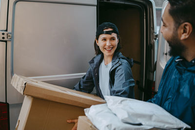 Smiling female delivery person talking with male coworker while carrying boxes near van