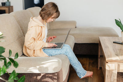 The schoolgirl sits on the couch and writes in a laptop. the child is learning using gadgets at home