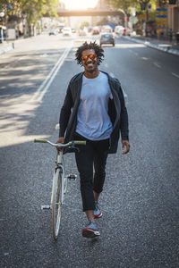 Man wearing sunglasses with bicycle walking on road in city