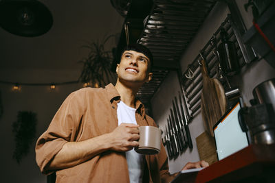 Low angle view of smiling male freelancer holding coffee cup with laptop on counter in kitchen at home office