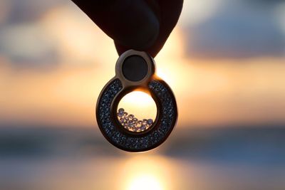 Cropped hand holding pendant against sky during sunset