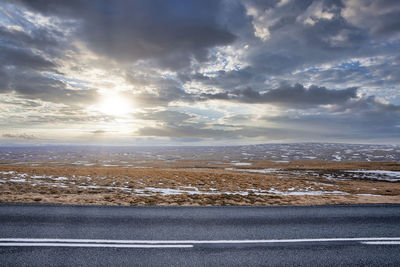 View of sun shining over road by snow covered landscape against cloudy sky