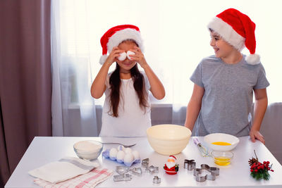 Brother and sister in red caps are having fun and preparing christmas cookies at a white table.