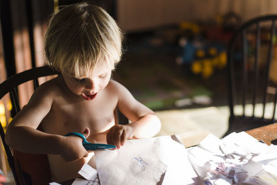 High angle view of shirtless boy cutting papers with scissors while sitting on chair at home